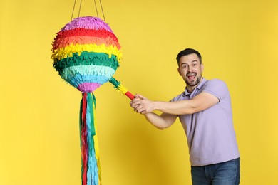 Emotional man hitting colorful pinata with stick on yellow background