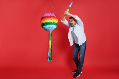 Emotional man hitting colorful pinata with stick on red background