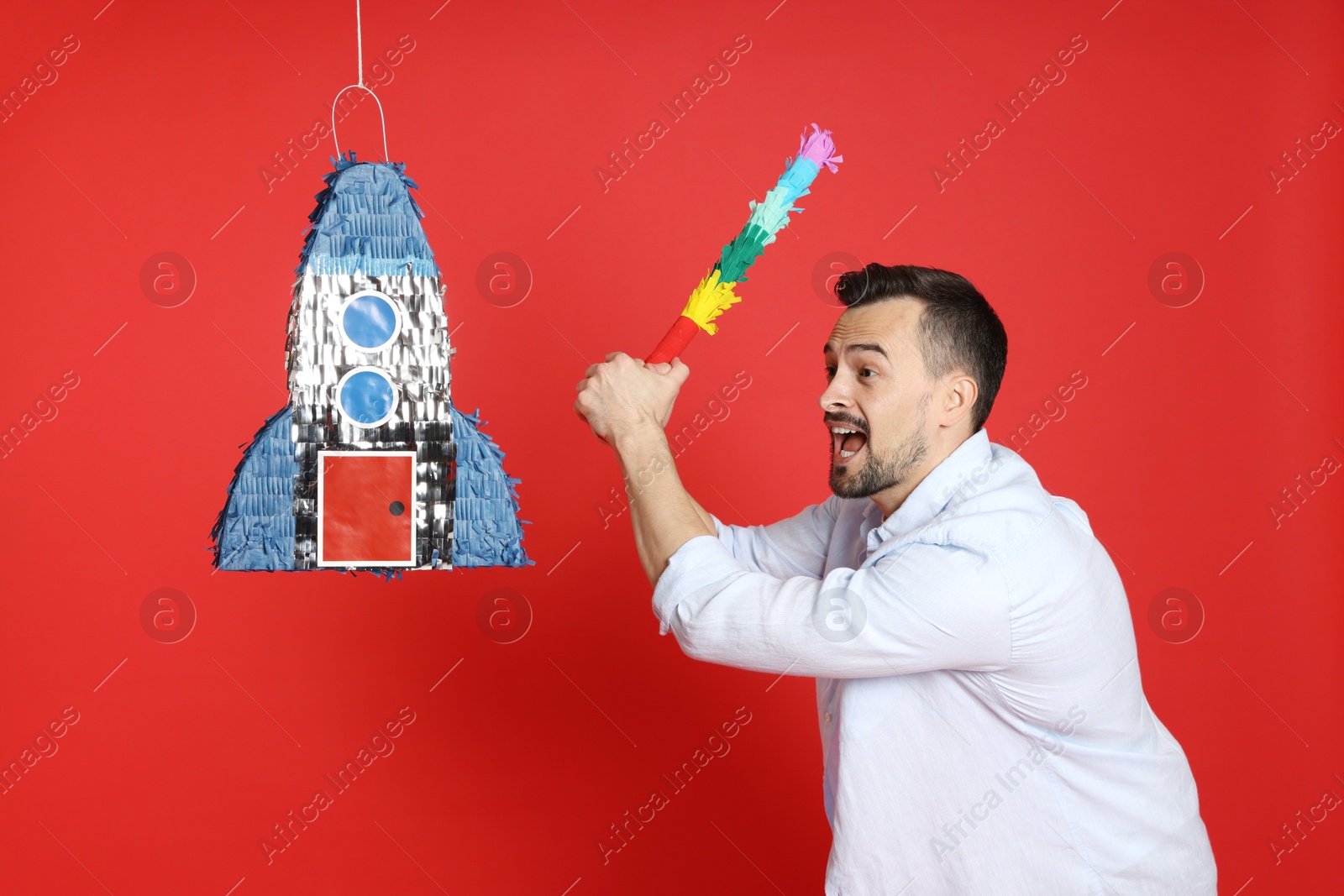 Photo of Emotional man hitting rocket shaped pinata with stick on red background