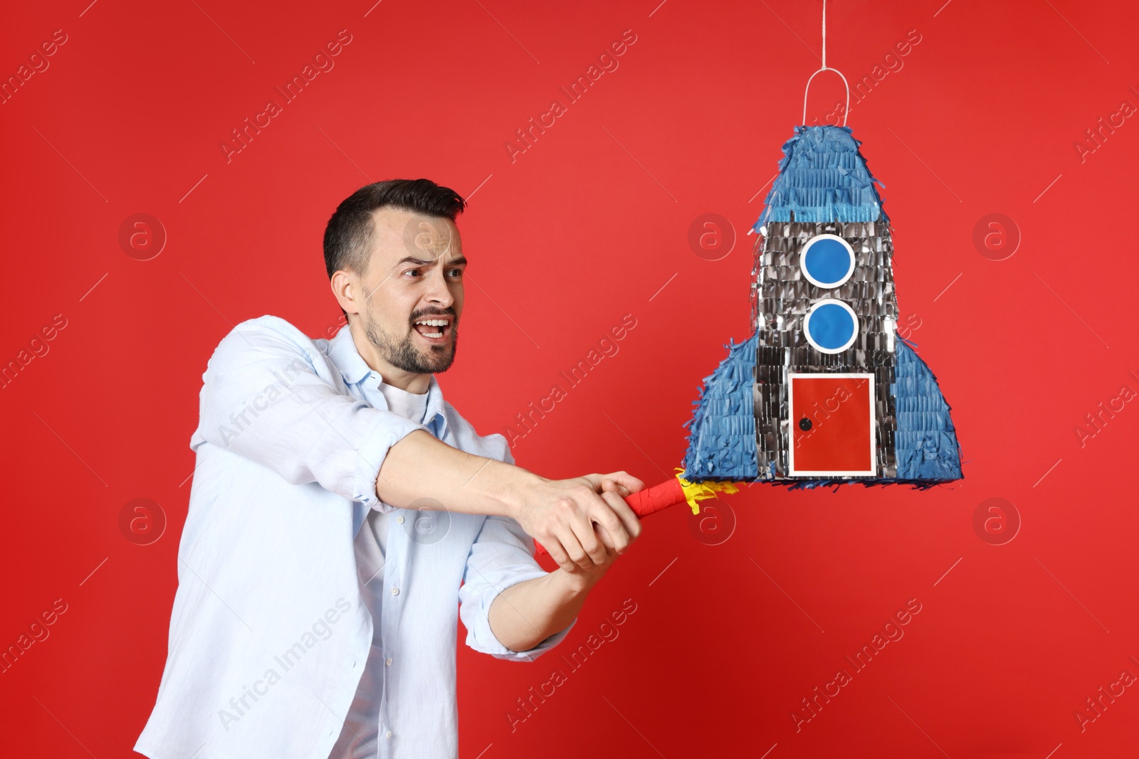 Photo of Emotional man hitting rocket shaped pinata with stick on red background