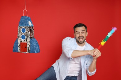 Emotional man hitting rocket shaped pinata with stick on red background