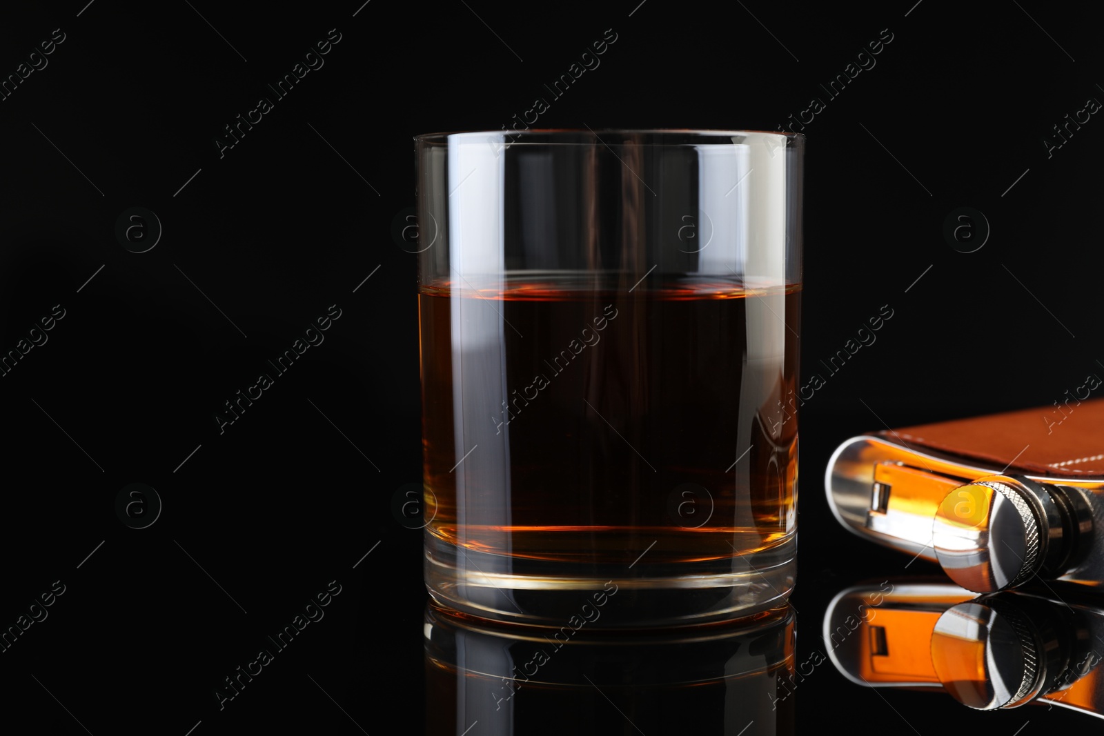 Photo of Hip flask and glass of whiskey on black mirror surface, closeup