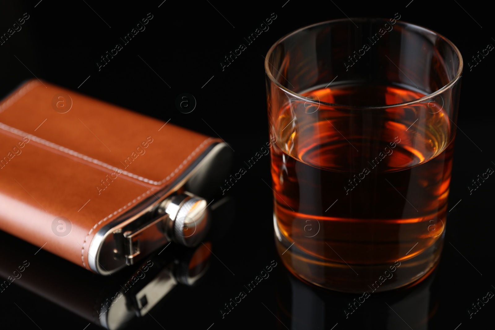 Photo of Hip flask and glass of whiskey on black mirror surface, closeup