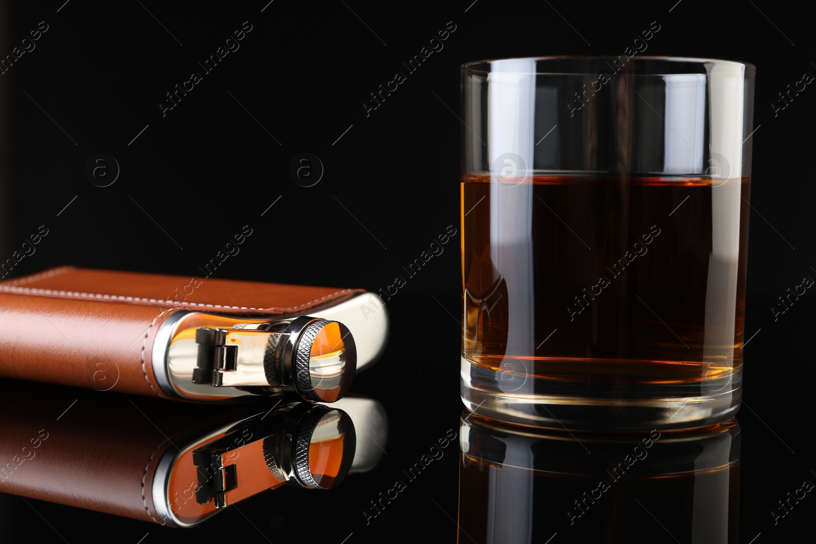 Photo of Hip flask and glass of whiskey on black mirror surface, closeup