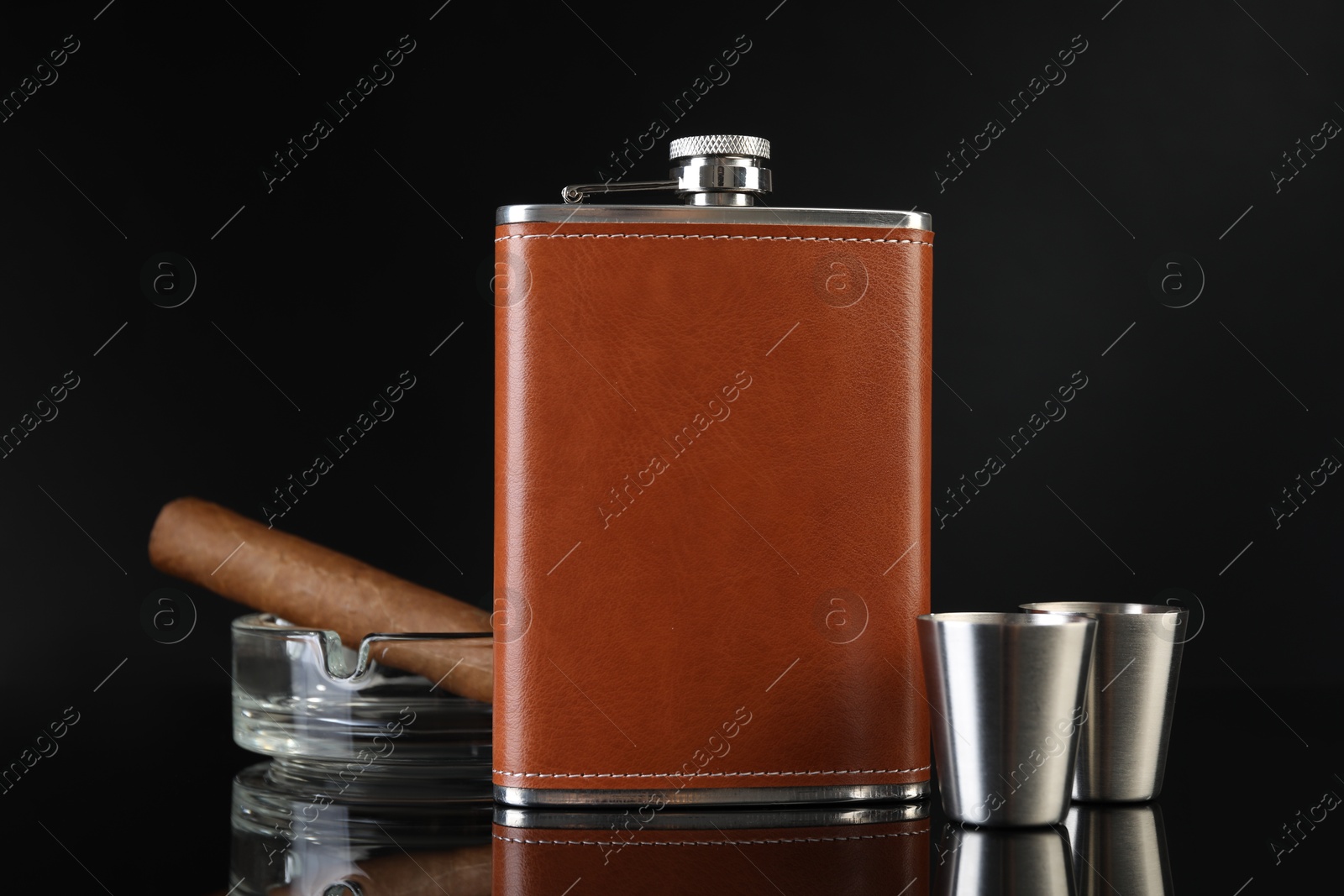 Photo of Hip flask, cups, cigar and ashtray on black mirror surface