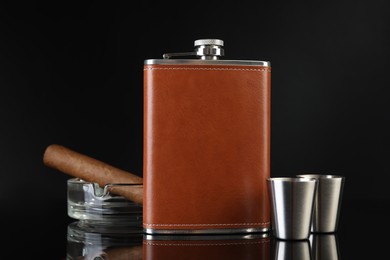 Photo of Hip flask, cups, cigar and ashtray on black mirror surface