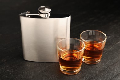 Photo of Hip flask and shot glasses of whiskey on black table, closeup