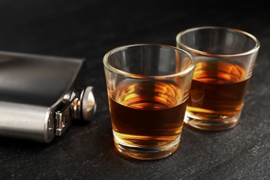 Photo of Hip flask and shot glasses of whiskey on black table, closeup