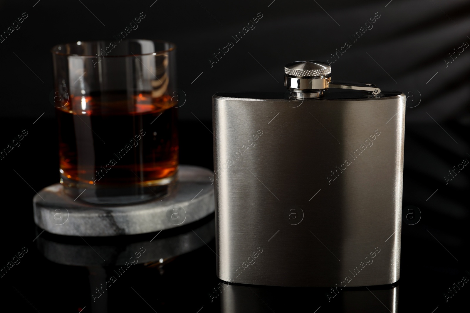 Photo of Hip flask and glass of whiskey on black mirror surface, closeup