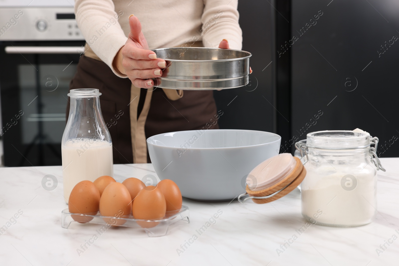 Photo of Making dough. Woman sifting flour into bowl at white table, closeup