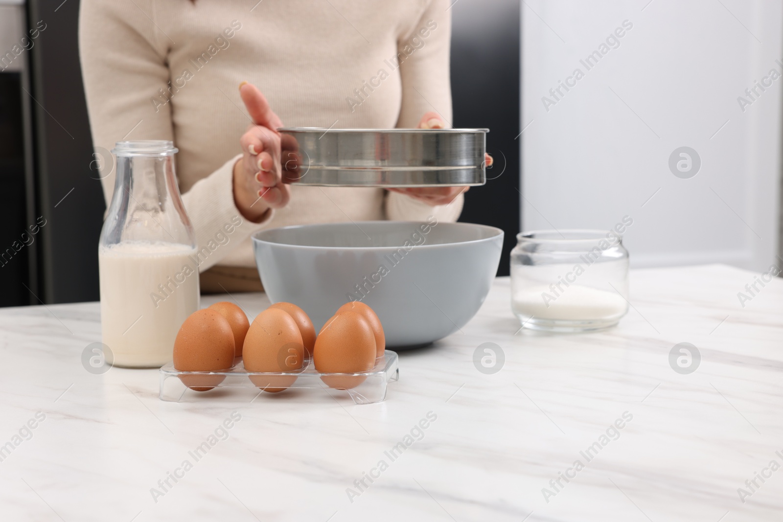 Photo of Making dough. Woman sifting flour into bowl at white table, closeup