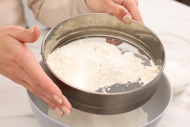 Photo of Making dough. Woman sifting flour into bowl at white table, closeup