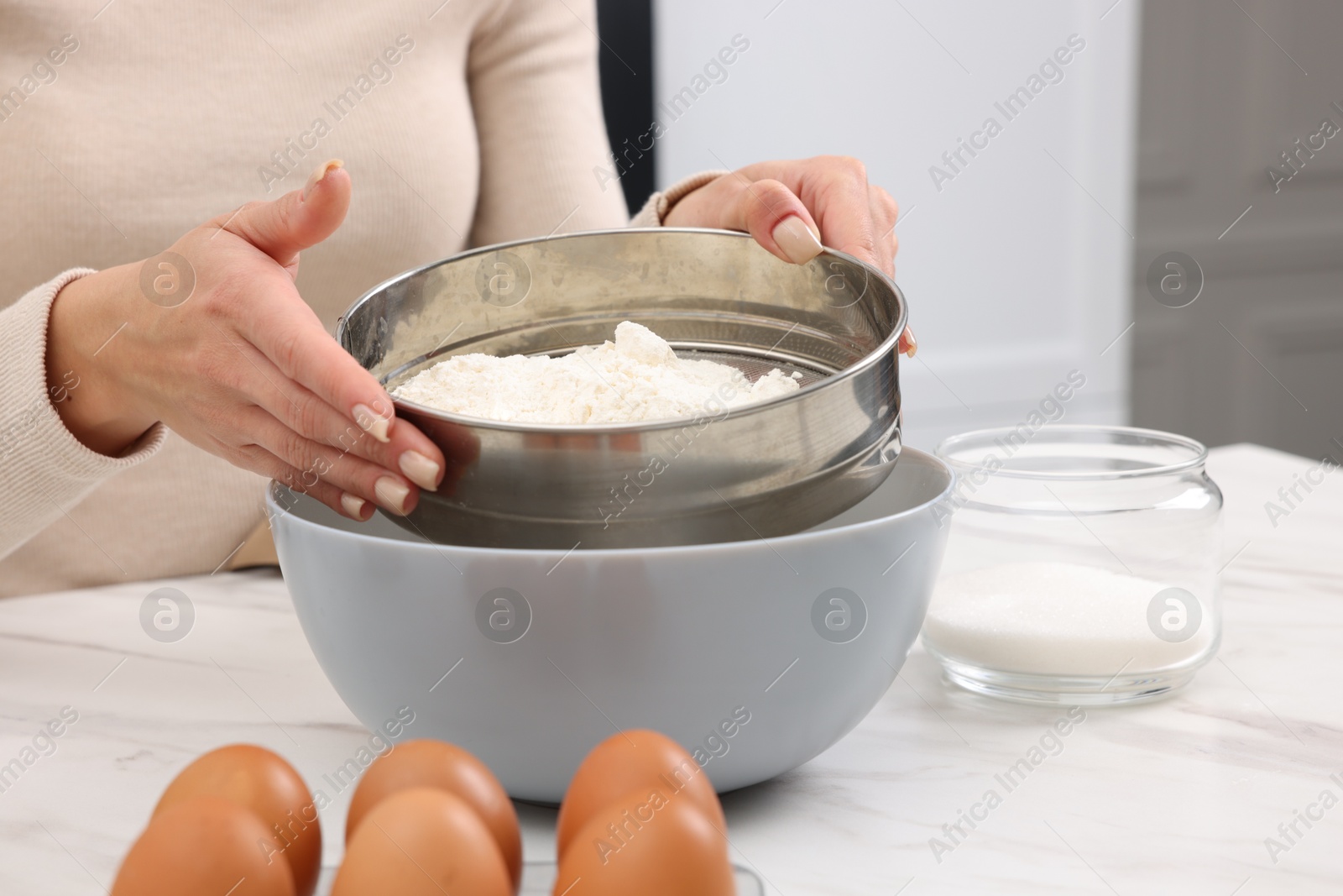 Photo of Making dough. Woman sifting flour into bowl at white table, closeup