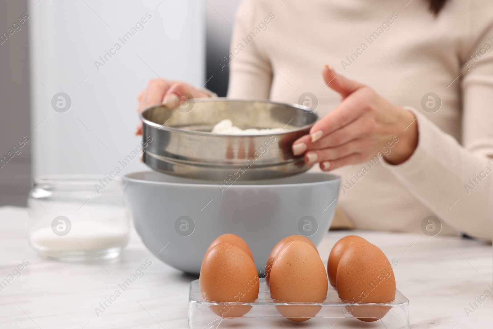 Photo of Making dough. Woman sifting flour into bowl at white table, closeup