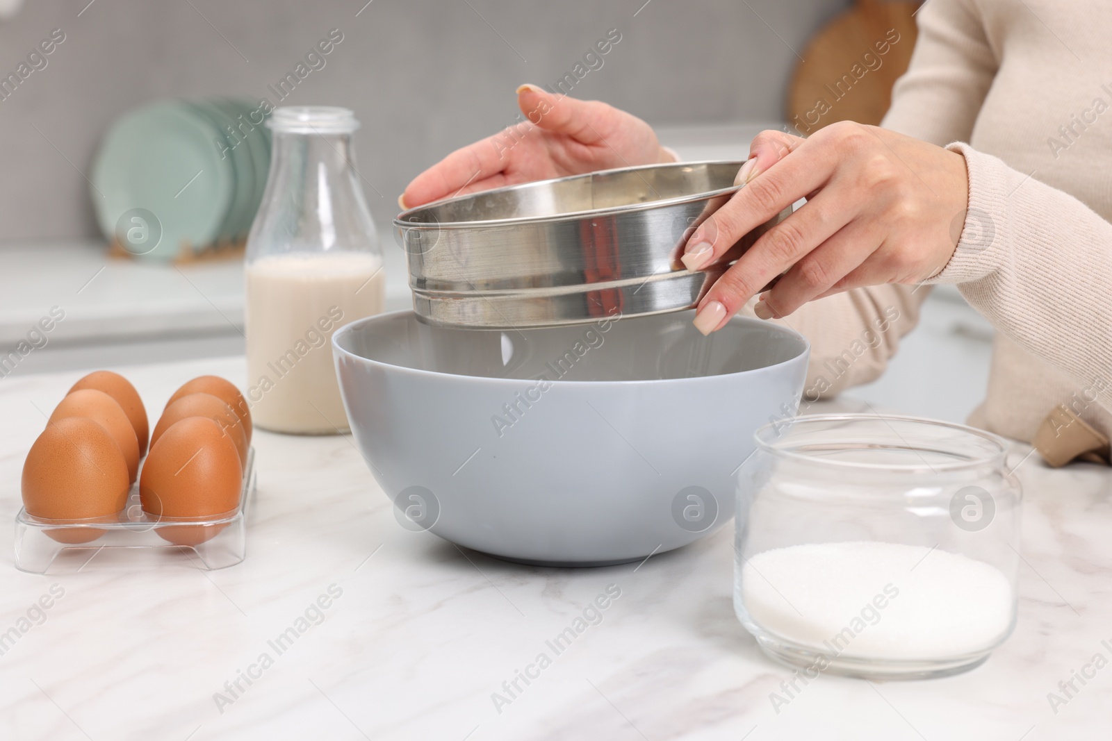 Photo of Making dough. Woman sifting flour into bowl at white table, closeup