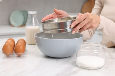 Photo of Making dough. Woman sifting flour into bowl at white table, closeup