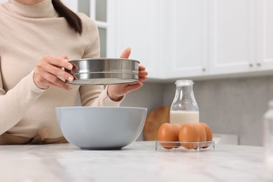 Photo of Making dough. Woman sifting flour into bowl at white table, closeup