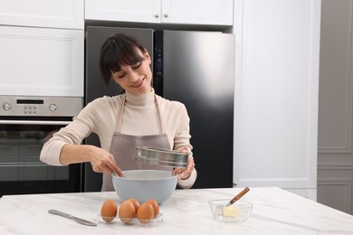 Woman sifting flour into bowl with dough at white marble table. Space for text