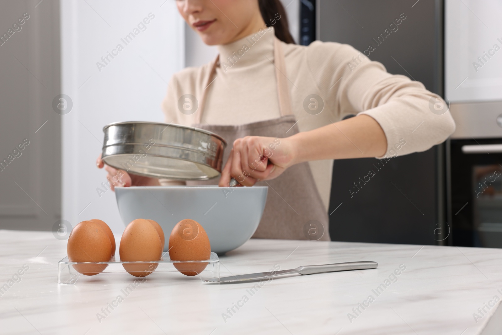 Photo of Woman sifting flour into bowl with dough at white marble table, closeup