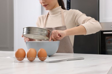 Woman sifting flour into bowl with dough at white marble table, closeup