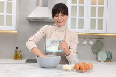 Woman sifting flour into bowl with dough at white marble table