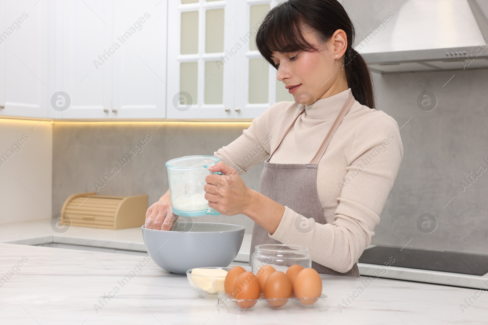 Photo of Woman sifting flour into bowl with dough at white marble table