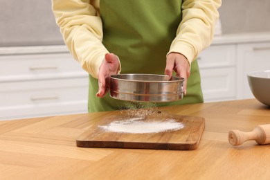 Woman sprinkling flour onto wooden board before rolling dough at table indoors, closeup