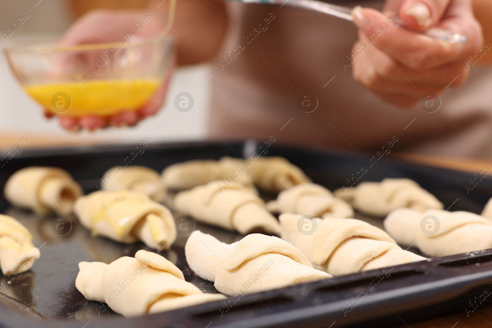 Photo of Woman brushing egg wash onto raw croissants at table indoors, closeup