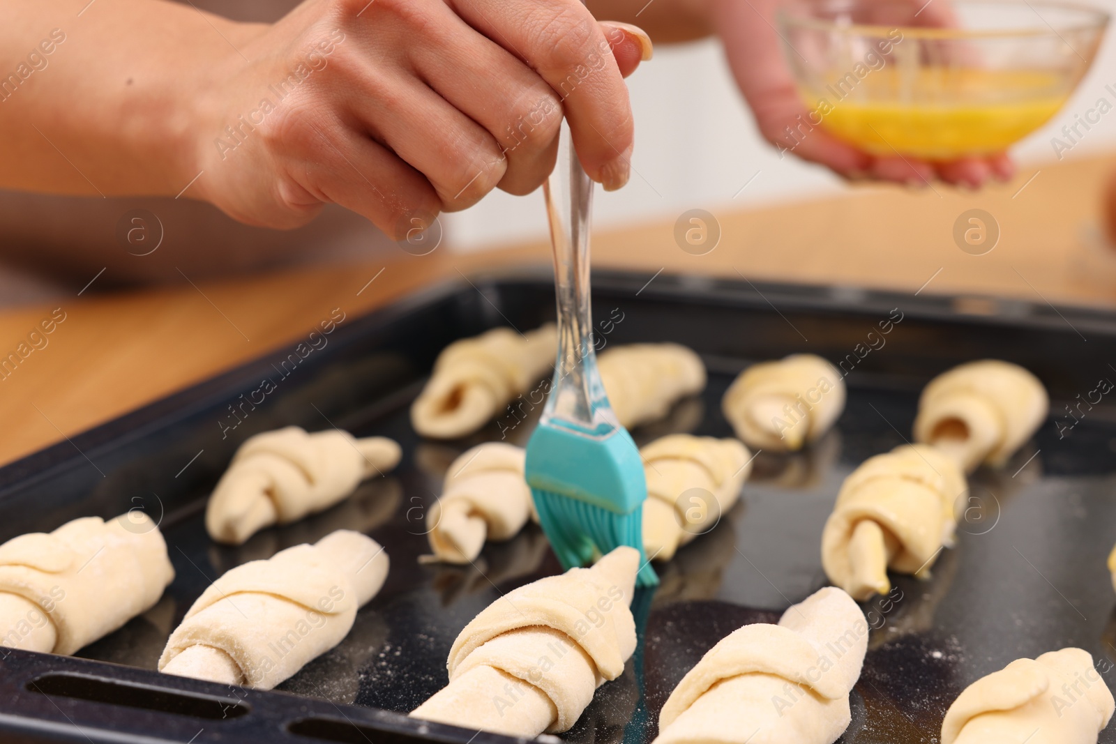 Photo of Woman brushing egg wash onto raw croissants at table indoors, closeup