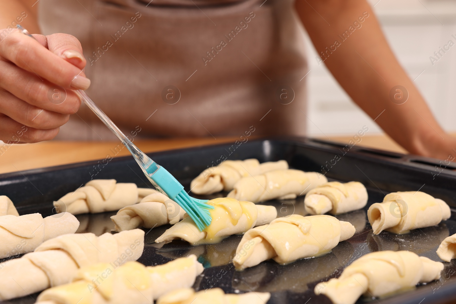 Photo of Woman brushing egg wash onto raw croissants at table indoors, closeup