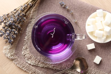 Aromatic lavender tea in glass cup, spoon, sugar cubes and bunch of dry flowers on wooden table, flat lay