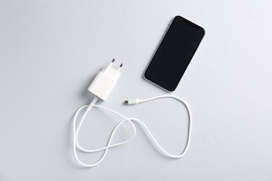 Photo of USB power adapter with charge cable and smartphone on white background, flat lay