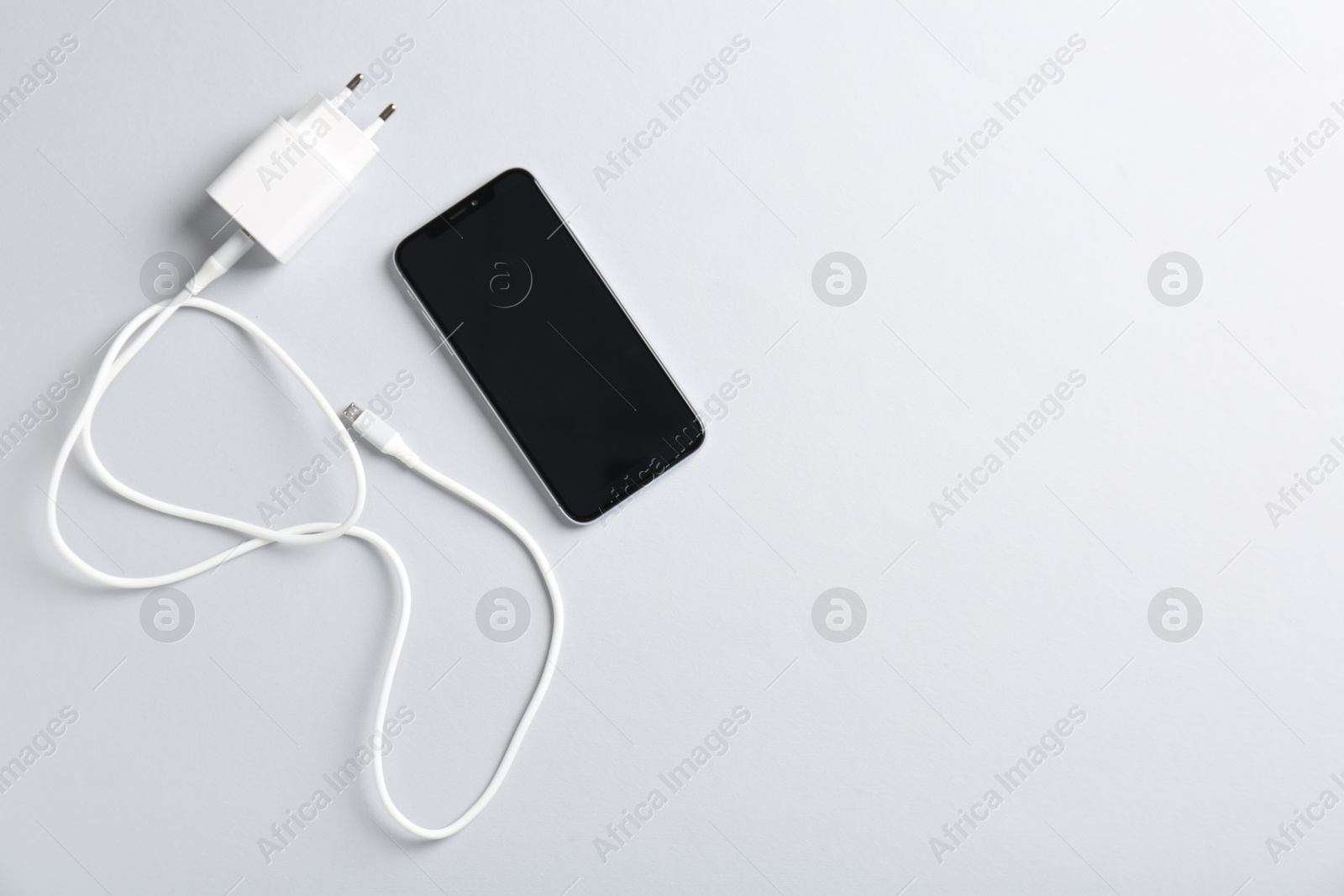 Photo of USB power adapter with charge cable and smartphone on white background, flat lay. Space for text