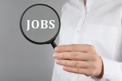 Image of Job search concept. Woman looking at word through magnifying glass on grey background, closeup