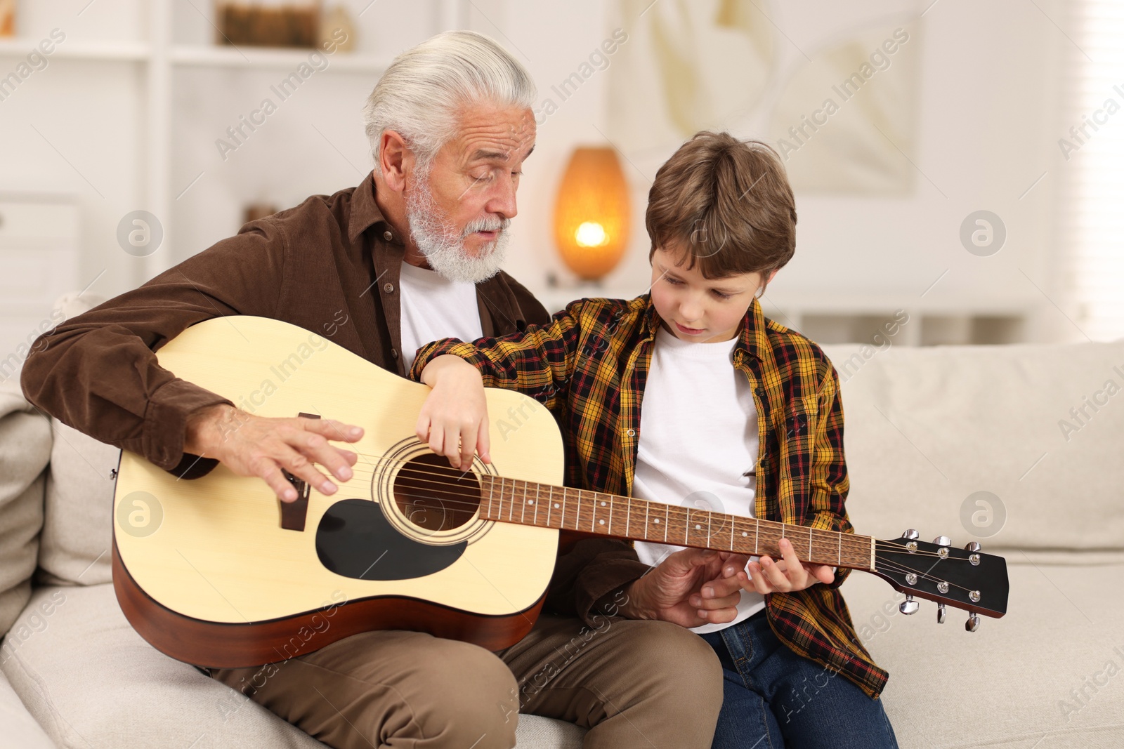 Photo of Grandpa teaching his grandson to play guitar on sofa at home