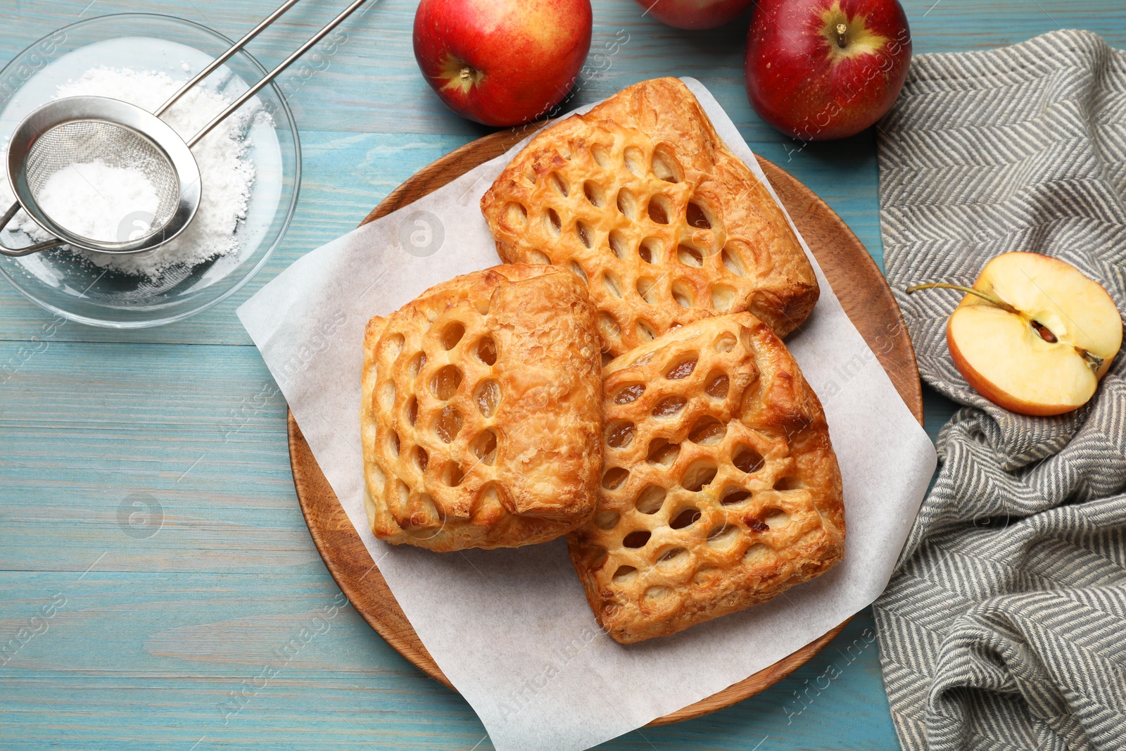 Photo of Delicious puff pastries, apples and powdered sugar on light blue wooden table, top view