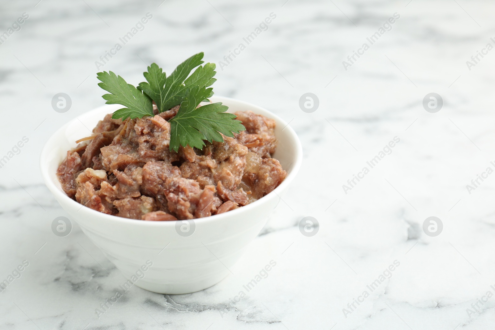 Photo of Canned meat with parsley in bowl on white marble table, closeup. Space for text