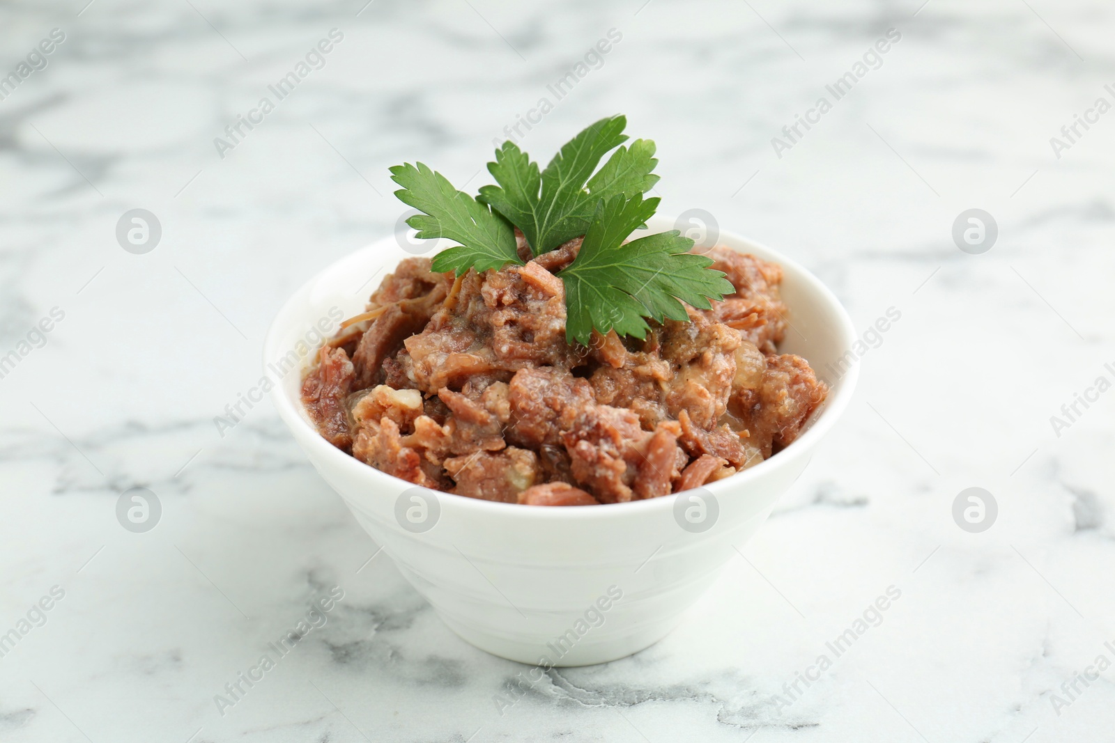 Photo of Canned meat with parsley in bowl on white marble table, closeup