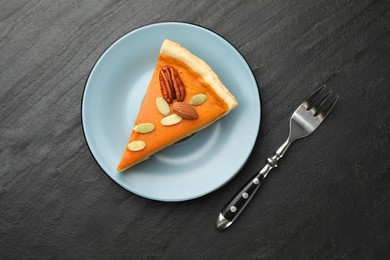 Photo of Piece of tasty homemade pumpkin pie with seeds, nuts and fork on black table, top view