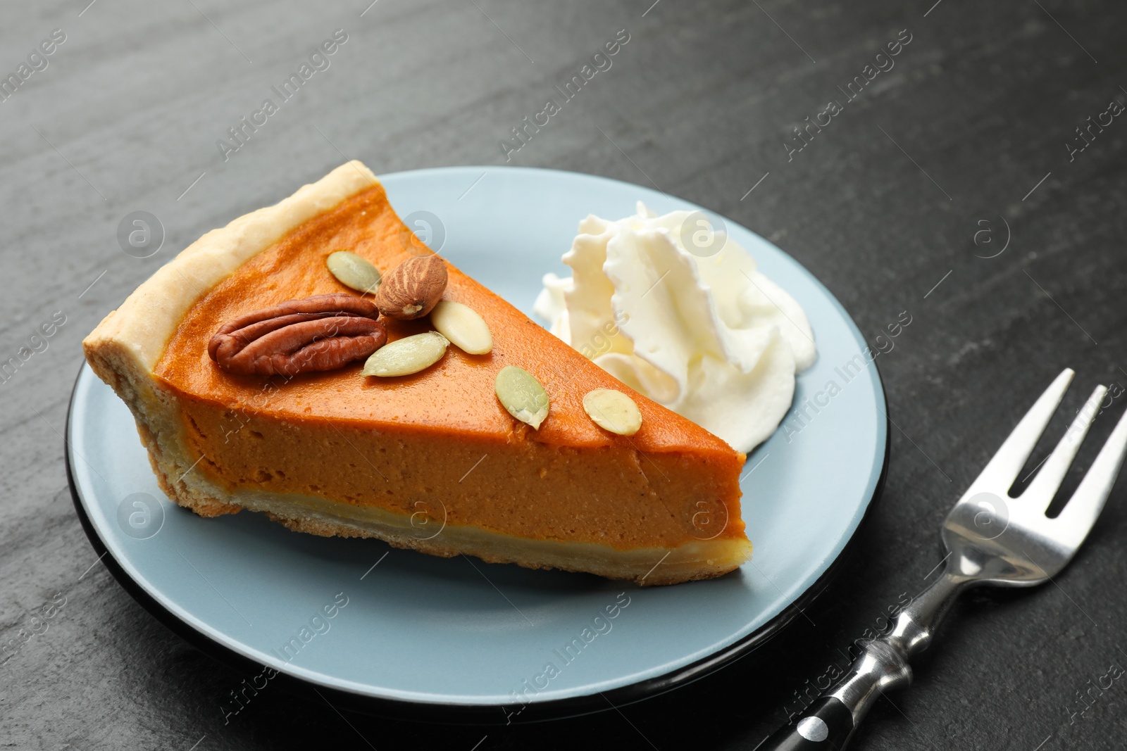 Photo of Piece of tasty homemade pumpkin pie with whipped cream, seeds, nuts and fork on black table, closeup