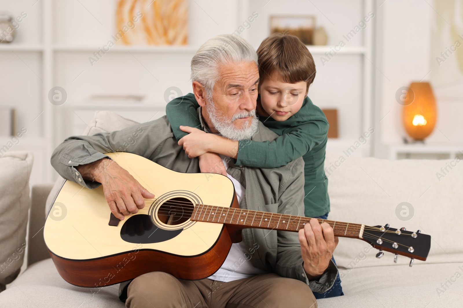 Photo of Grandpa teaching his grandson to play guitar on sofa at home