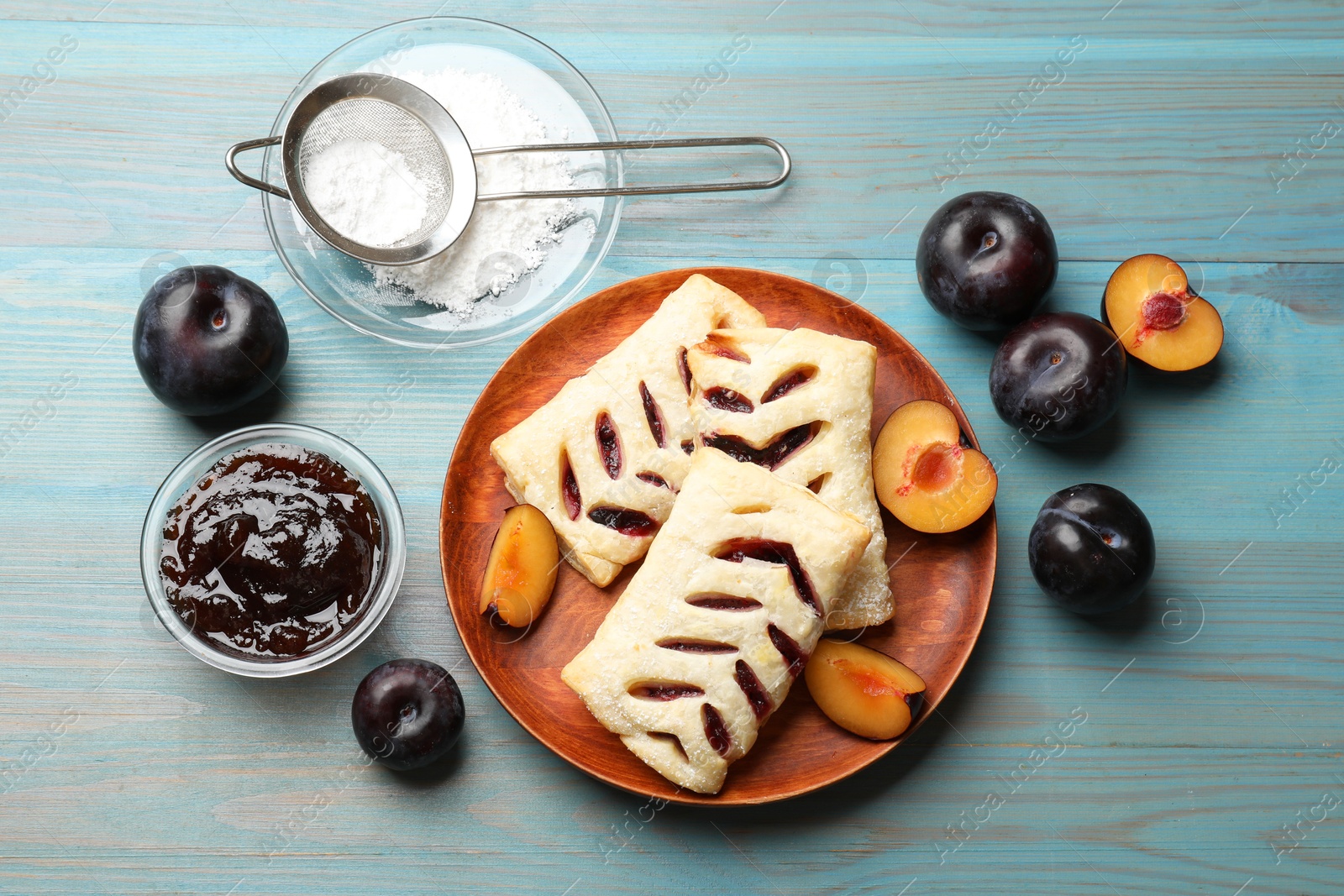 Photo of Delicious puff pastries, plums, jam and powdered sugar on light blue wooden table, top view