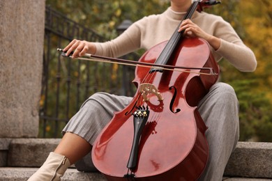 Photo of Young woman playing cello on stairs outdoors, closeup. Classic musical instrument