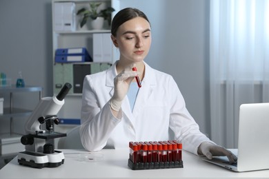 Laboratory testing. Doctor holding test tube with blood sample while working on laptop at table indoors