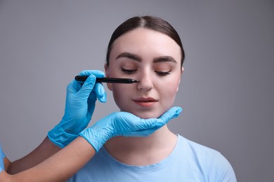 Photo of Doctor drawing marks on patient's nose for plastic surgery operation against grey background, closeup