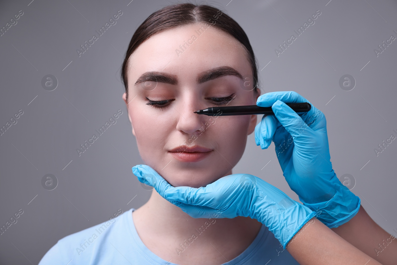 Photo of Doctor drawing marks on patient's nose for plastic surgery operation against grey background, closeup