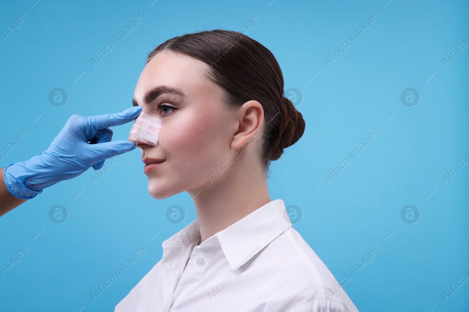 Photo of Doctor checking patient's nose after plastic surgery operation on light blue background, closeup