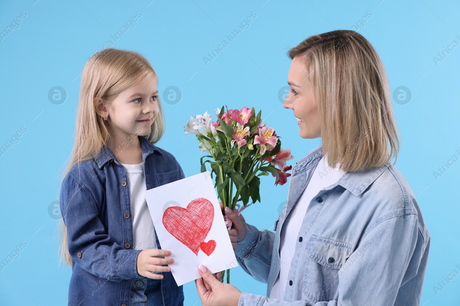 Photo of Little daughter congratulating her mom with bouquet of alstroemeria flowers and greeting card on light blue background. Happy Mother's Day