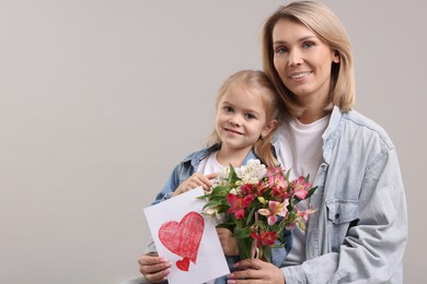 Photo of Happy woman with her daughter, bouquet of alstroemeria flowers and greeting card on gray background, space for text. Mother's Day celebration