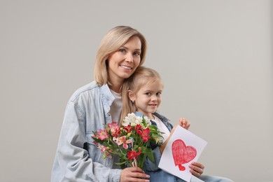 Photo of Happy woman with her daughter, bouquet of alstroemeria flowers and greeting card on gray background. Mother's Day celebration